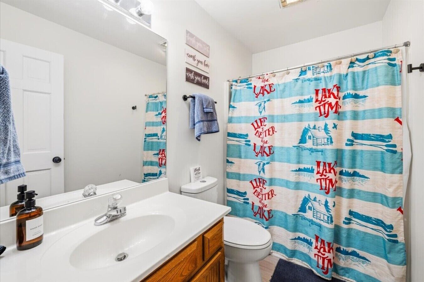 Kylpyhuoneessa on valkoinen pesuallas, puinen kaappi ja wc-istuin. Siniraidallinen, valkoisella pohjalla oleva suihkuverho, jossa on teksti ”LAKE TIM”, on näkyvissä.
