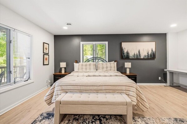 A bedroom with a large bed, two bedside tables, a window with blinds, a painting on the wall, and a rug.