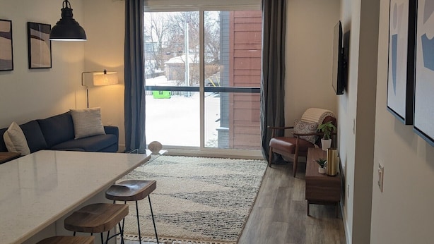 A modern kitchen with a white countertop and bar stools, a dining area with a large window, and a living space with a sofa and a coffee table. - Luxury 1 Bedroom Downtown Traverse City (Traverse City)