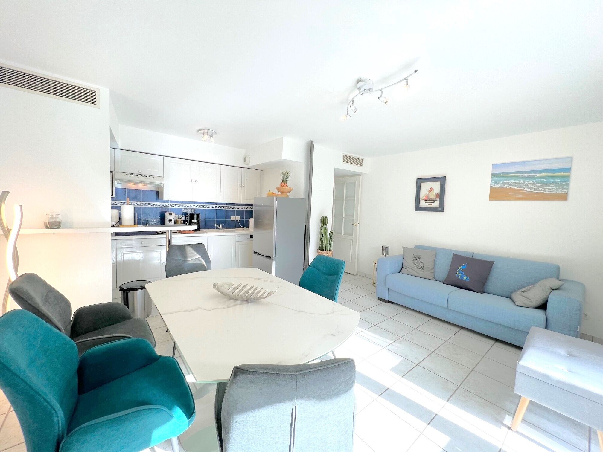 A modern kitchen with white cabinets, a dining area with a white table and teal chairs, and a light blue sofa in a bright living room.