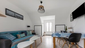 Un salon moderne avec un canapé, une table basse et une table à manger. Un téléviseur est fixé au mur et une fenêtre laisse entrer la lumière naturelle.