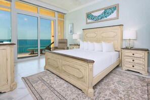 A bedroom with a large bed, two bedside tables, a chair, a view through a sliding door to a balcony, and a painting on the wall.