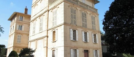 Um edifício de vários andares com fachada simétrica, janelas grandes com persianas e uma entrada central.