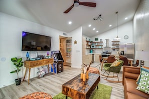 Een moderne woonkamer met een flatscreen televisie, een houten salontafel, een groene bank en een eethoek met stoelen, met een keuken op de achtergrond.