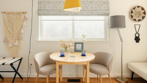 Ett rum med ett runt träbord, två beige stolar, en gul hänglampa, en golvlampa, en vävkonst i makramé och ett fönster med utsikt.