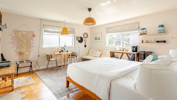 Ett mysigt hotellrum med en stor säng, ett skrivbord, en stol och ett litet bord. Det finns två fönster med gardiner, en vägghylla med diverse föremål och en hängande lampa.