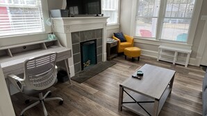 A modern living room with a fireplace, a TV mounted above it, a desk with a chair, and a yellow armchair.