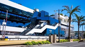 A modern building with a glass facade, a prominent staircase, and palm trees in the foreground.