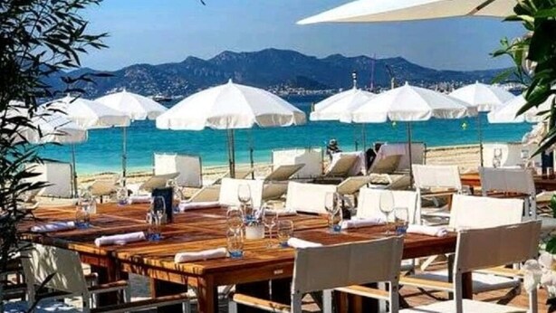 A beachside restaurant with outdoor seating, white umbrellas, and a view of the sea and mountains.