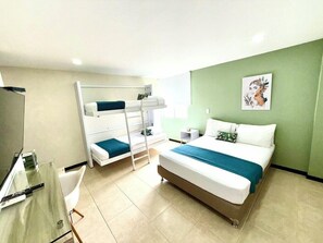 Quarto de hotel com beliches, uma cama grande, escrivaninha com cadeira, uma mesinha e televisão fixada na parede.