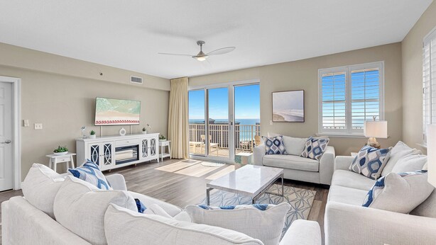 A spacious living room with a large flat-screen TV, a ceiling fan, and a balcony with a view of the ocean. - Ariel Dunes II 1610 - Corner Unit, Gulf Views, Pool, Fitness, Onsite Dining! (Miramar Beach)