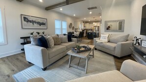 Ein geräumiges Wohnzimmer mit einer großen Couch, einem Couchtisch und einem Deckenventilator. Im Hintergrund ist eine Küche mit modernen Geräten und ein Essbereich zu sehen.