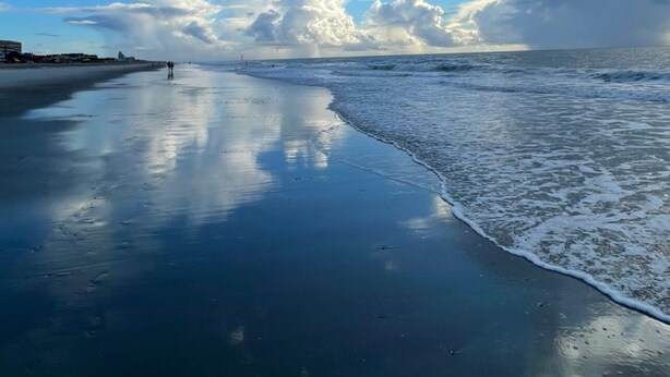 Ein Strand mit feuchtem Sand, der Himmel und Wolken widerspiegelt.