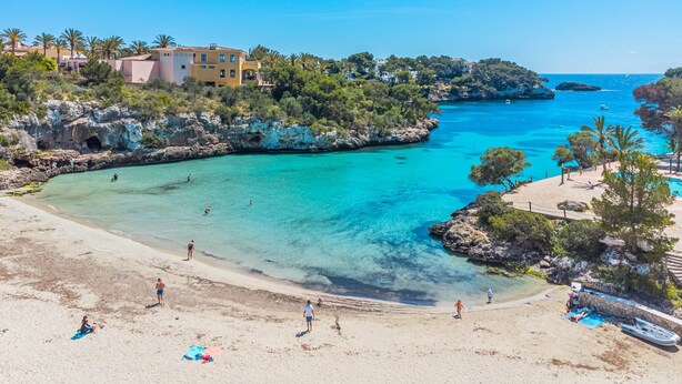 Ein Strand mit klarem türkisblauem Wasser, sandigem Ufer und Gebäuden im Hintergrund.
