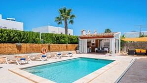 Ein rechteckiger Pool mit klarem, blauem Wasser, umrahmt von beige farbenen Fliesen. Liegestühle mit blauen Kissen und ein gelber Sonnenschirm sind vorhanden. Im Hintergrund sind eine Palme und ein Heckenstreifen zu sehen.