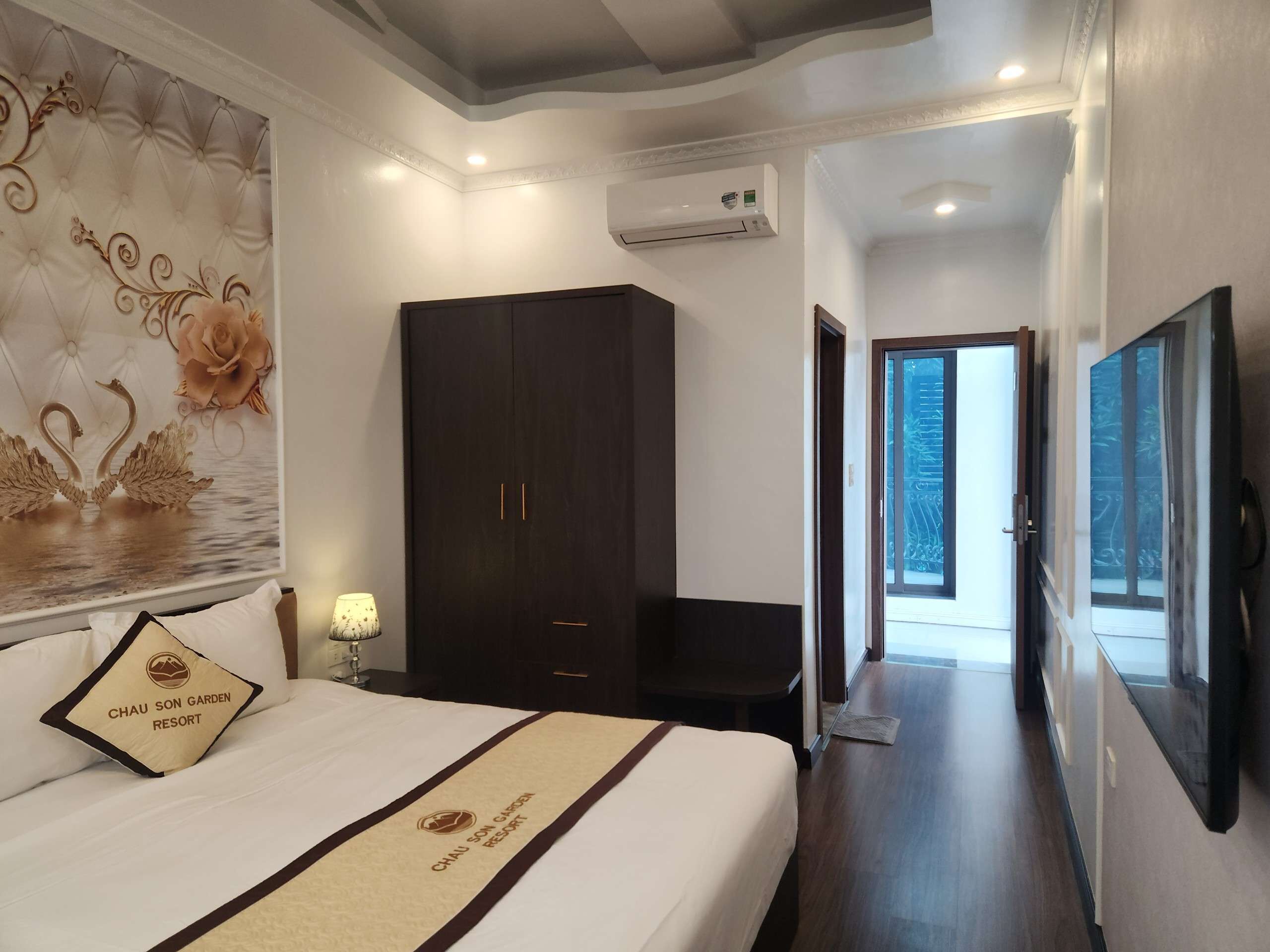 A hotel room with a bed, a wardrobe, a television, and a hallway leading to a balcony.