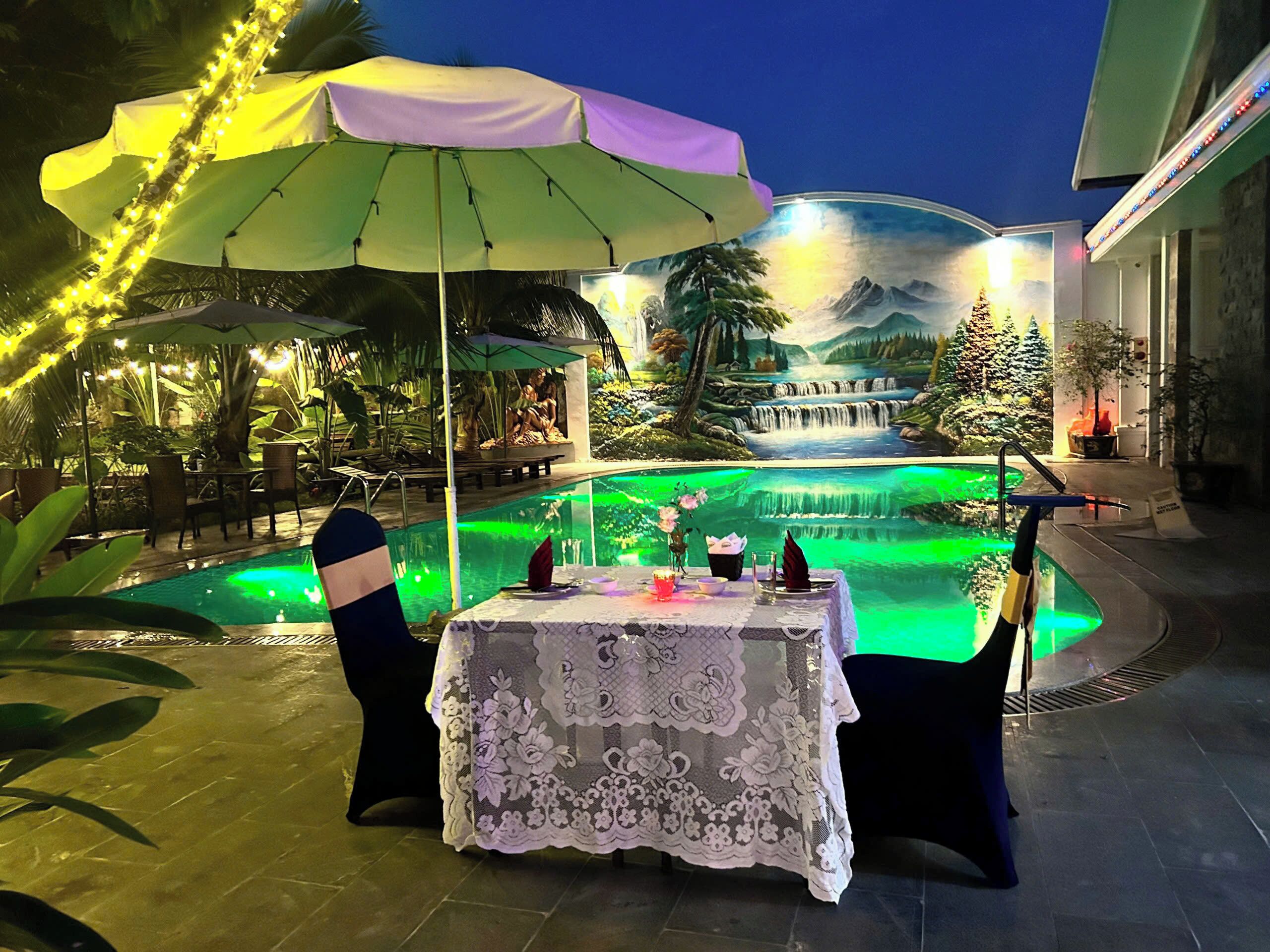 Uma área de refeições ao ar livre com uma mesa posta para dois, piscina com iluminação verde e uma cascata ao fundo.