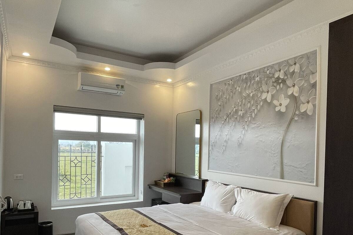 A bedroom with a large bed, a window with a view, a wall-mounted air conditioner, and a decorative wall piece.