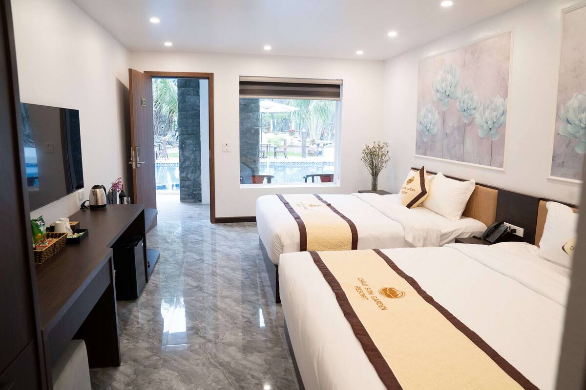 A hotel room with two beds, a desk, a TV, and a large window with a view of the outdoor area.