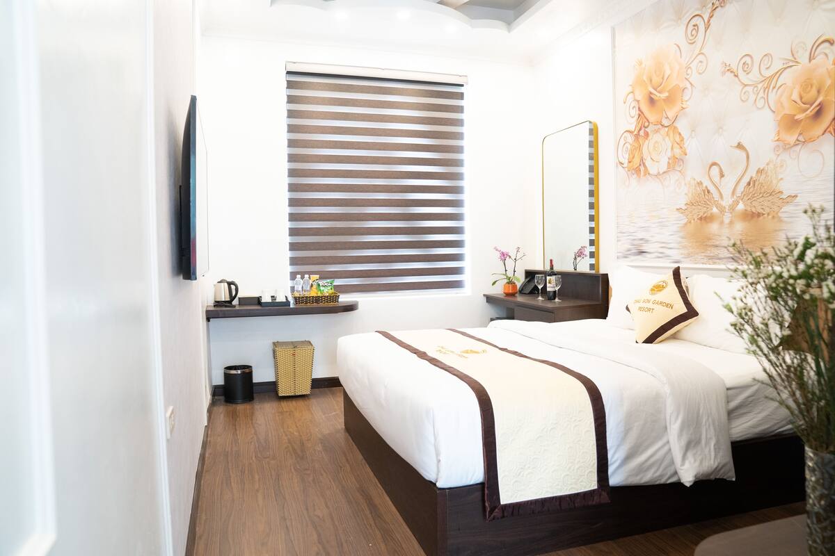 A hotel room with a large bed, a bedside table with a lamp, a window with blinds, a wall-mounted TV, and a decorative wall piece.