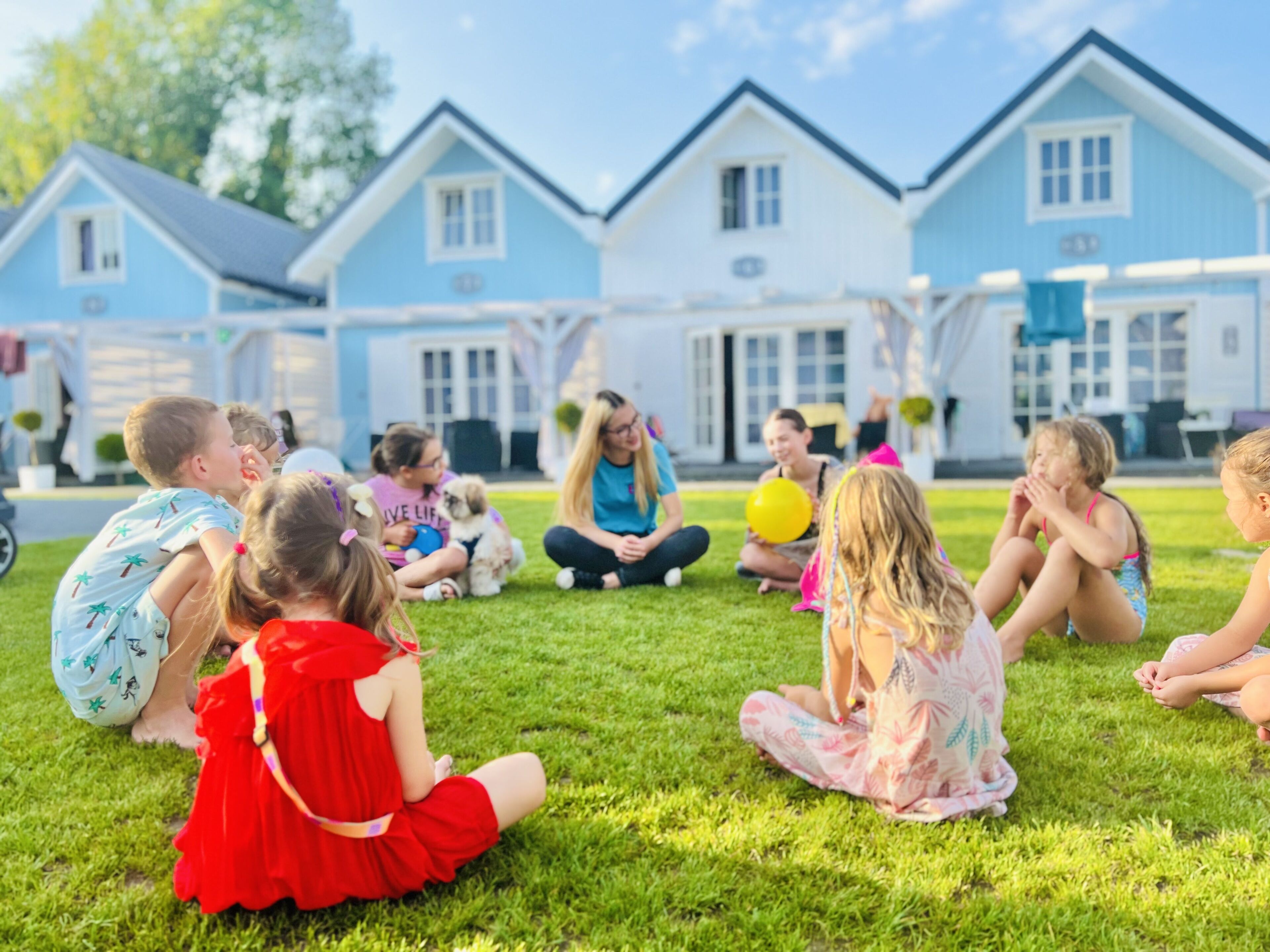 A group of children sitting on grass in front of a row of blue houses with white trim.