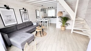 Um interior moderno com um sofá modular cinzento, uma mesa redonda com um vaso, cadeiras de cozinha brancas, uma área de cozinha e uma escadaria.