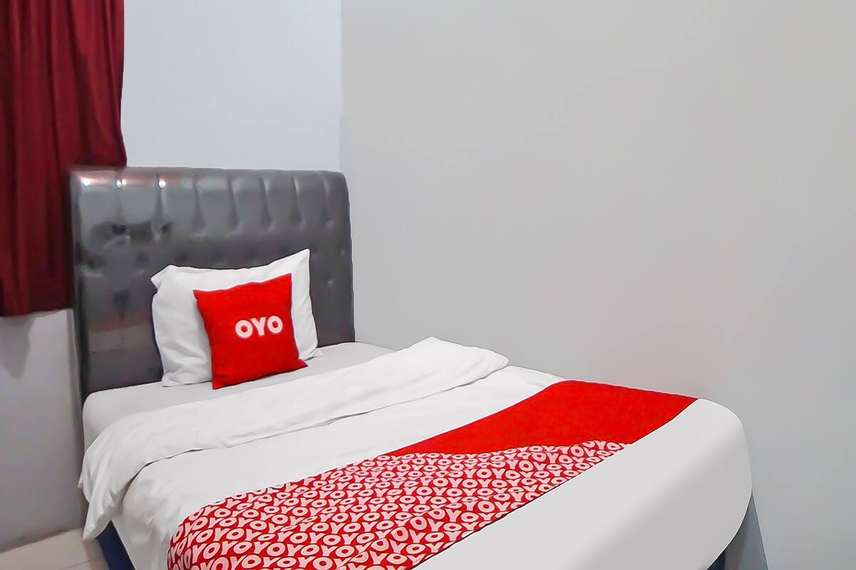Hotellihuone, jossa on harmaalla kankaalla verhoiltu sängynpääty, valkoiset vuodevaatteet ja punainen, kuvioitu heittopeitto.