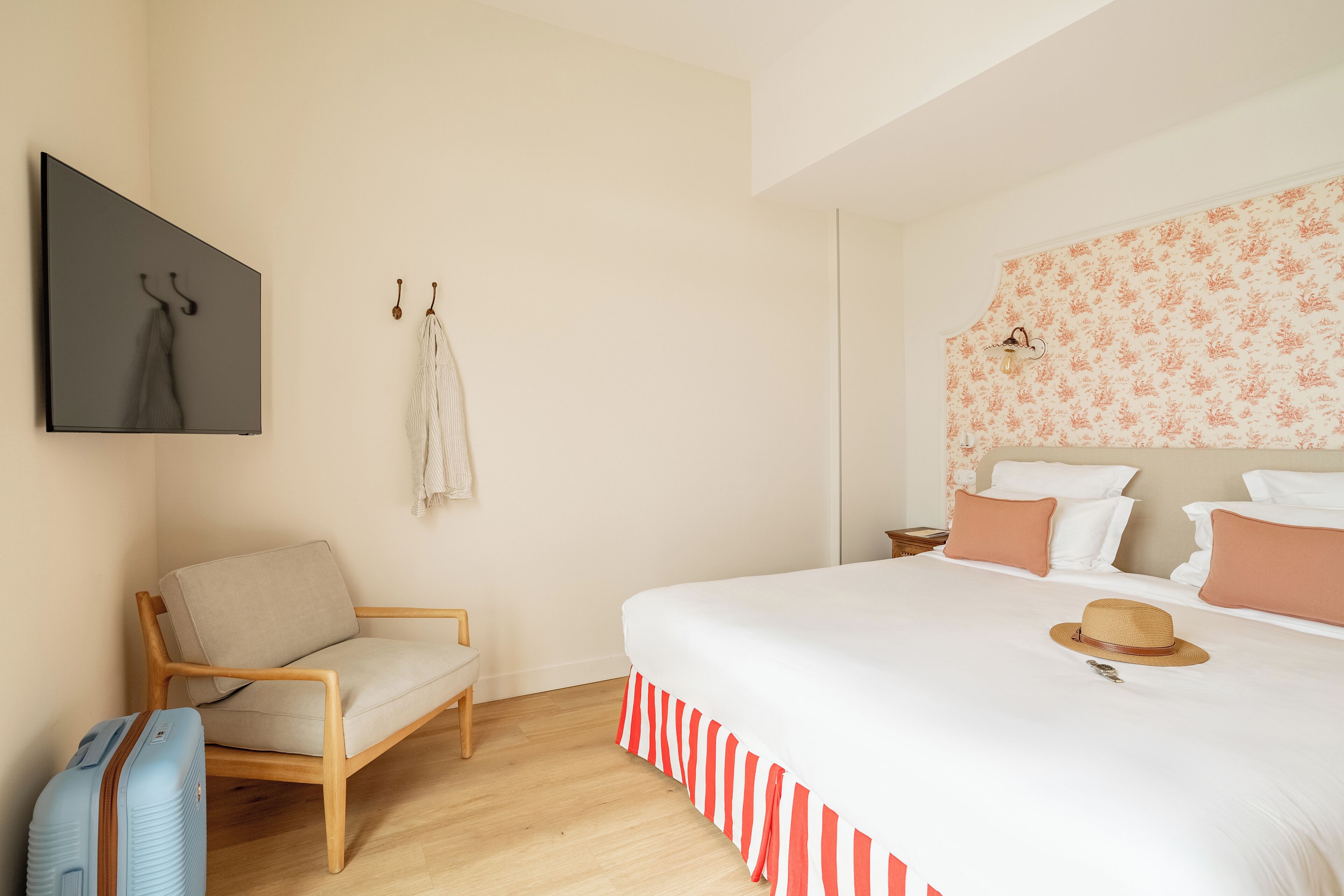 Un dormitorio con cama, una silla, una maleta y un televisor instalado en la pared.