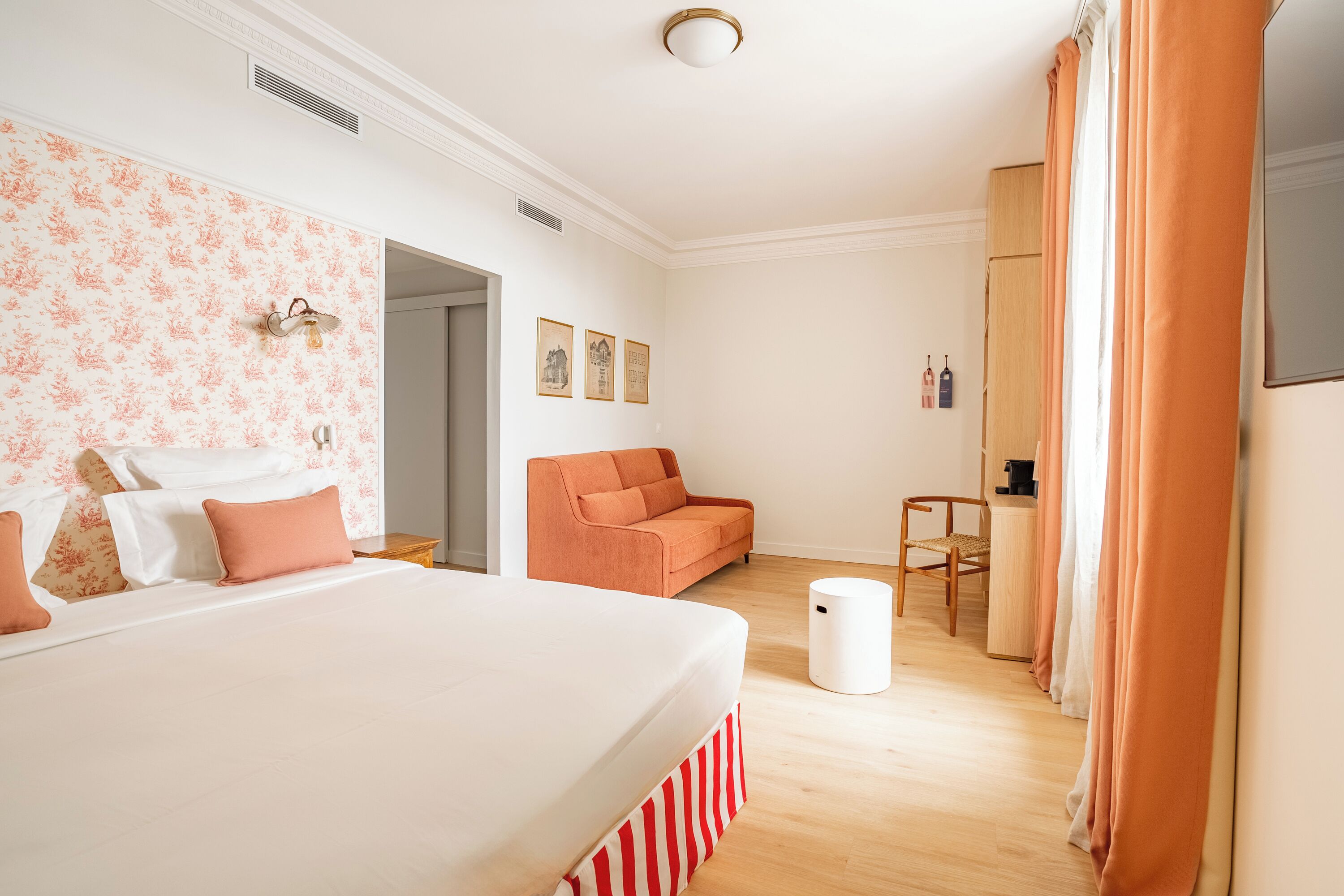 Un dormitorio con una cama grande, un sofá, una mesita y una silla. Hay cuadros en la pared y una ventana con cortinas.