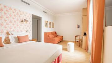 Un dormitorio con una cama grande, un sofá, una mesita y una silla. Hay cuadros en la pared y una ventana con cortinas.