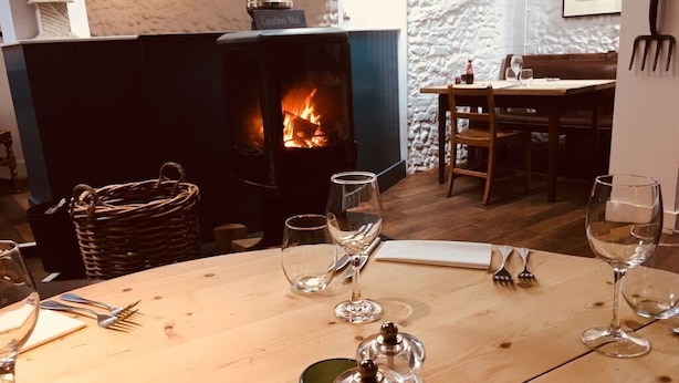 A cozy dining area with a wooden table set for two, a fireplace with a visible fire, and a rustic stone wall in the background. - The Kings Head (Holt)