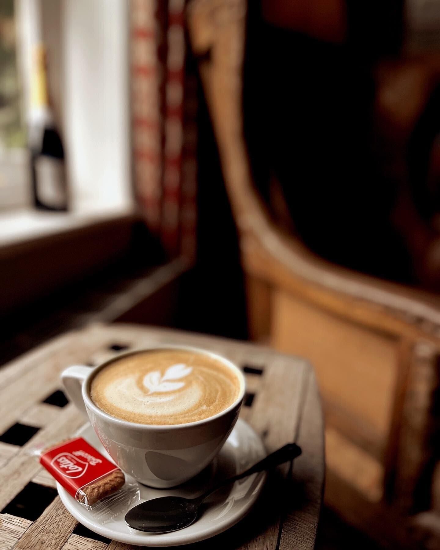 Una taza de café con arte de latte sobre un plato, una cuchara y una galleta sobre una mesa.
