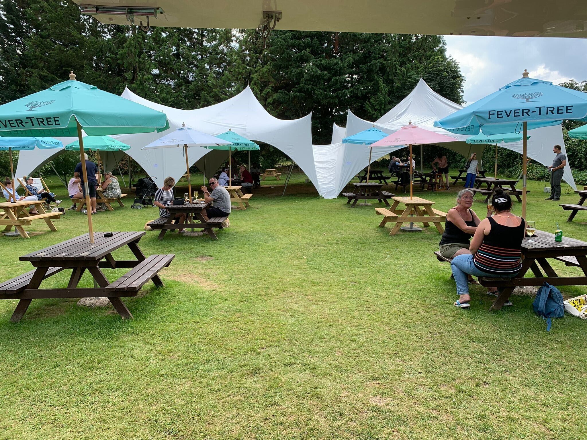 An outdoor picnic area with multiple tables and benches under umbrellas, surrounded by trees.