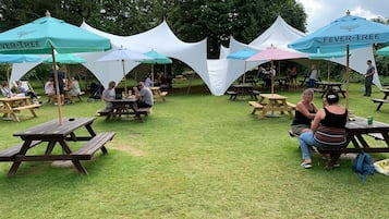 An outdoor picnic area with multiple tables and benches under umbrellas, surrounded by trees.