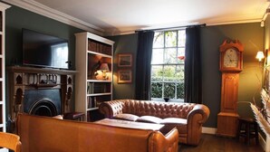 A cozy living room with a leather sofa, a fireplace, a bookshelf, and a large window with curtains. - The Kings Head (Holt)