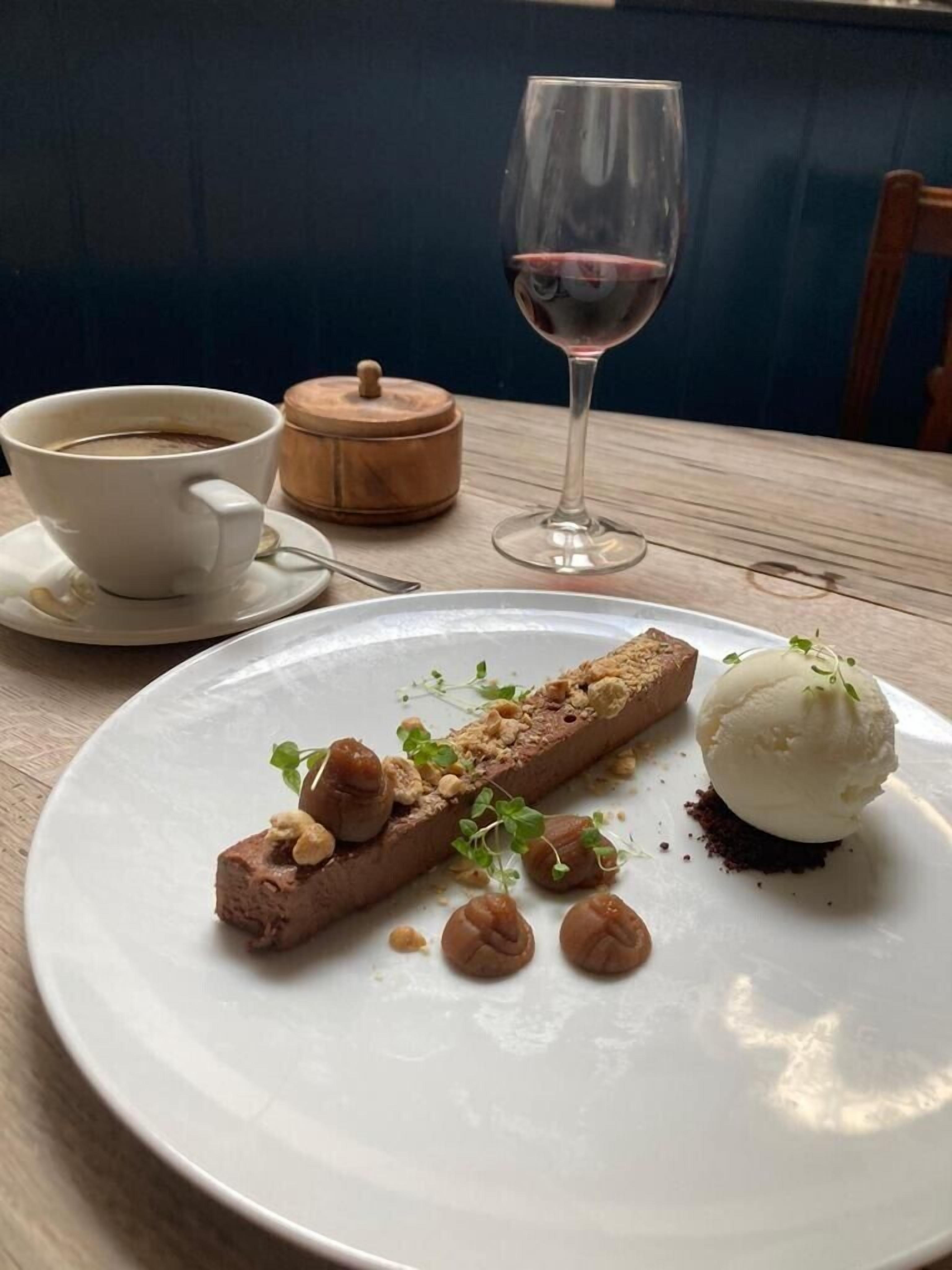 A dessert plate with a chocolate mousse and a scoop of ice cream, a cup of coffee, and a wine glass on a wooden table.