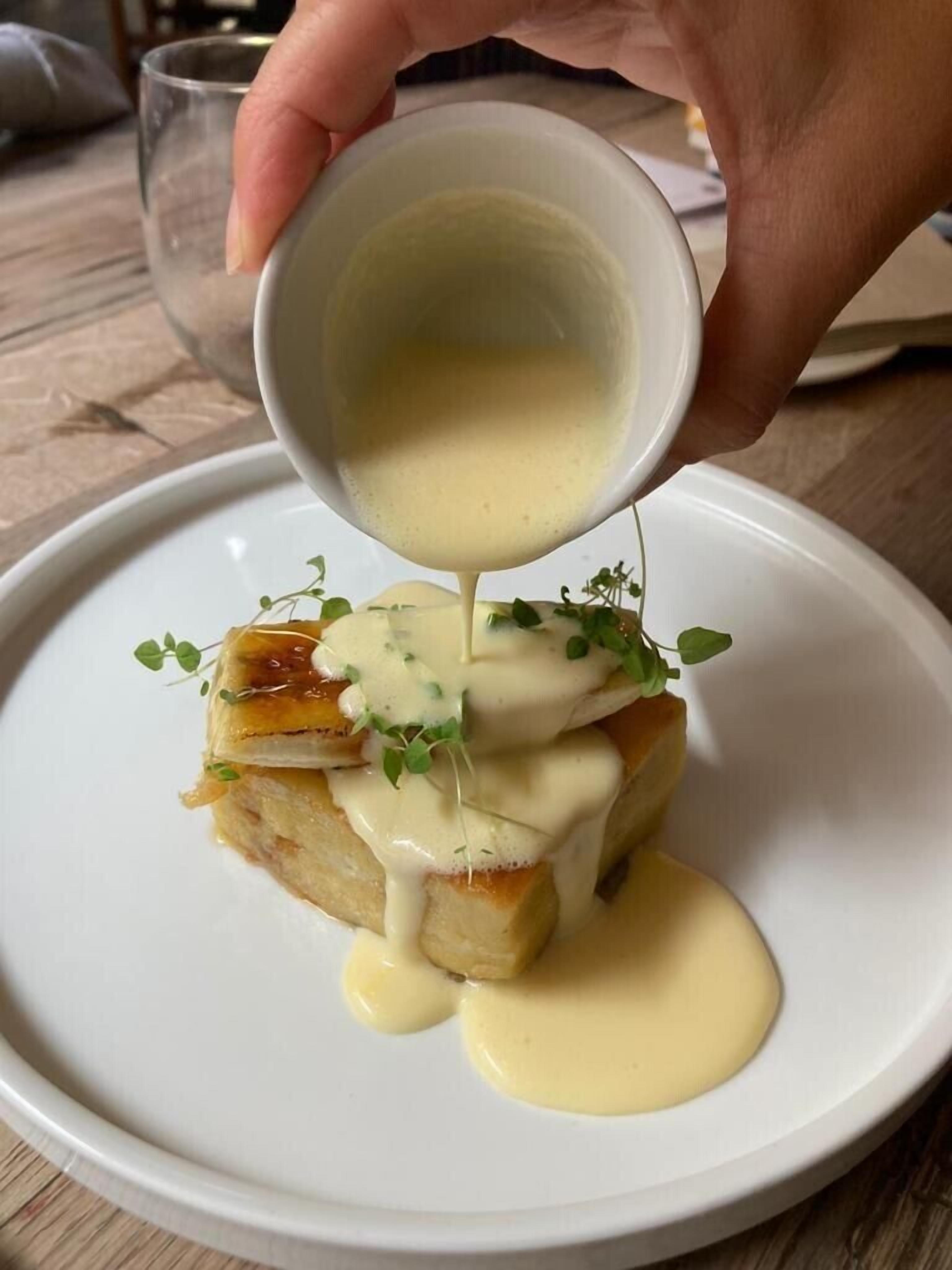 A hand pouring sauce over a dish with a piece of bread and greens.