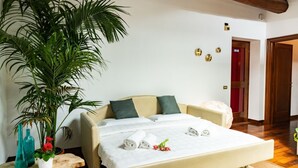 Quarto de hotel com uma cama, uma planta grande em vaso e uma mesinha de cabeceira de madeira com dois vasos.