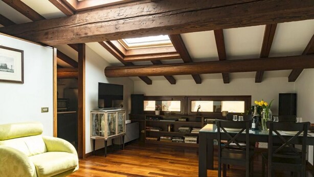 Um quarto amplo com vigas de madeira, um claraboia, área de refeições com mesa e cadeiras, e uma poltrona verde.