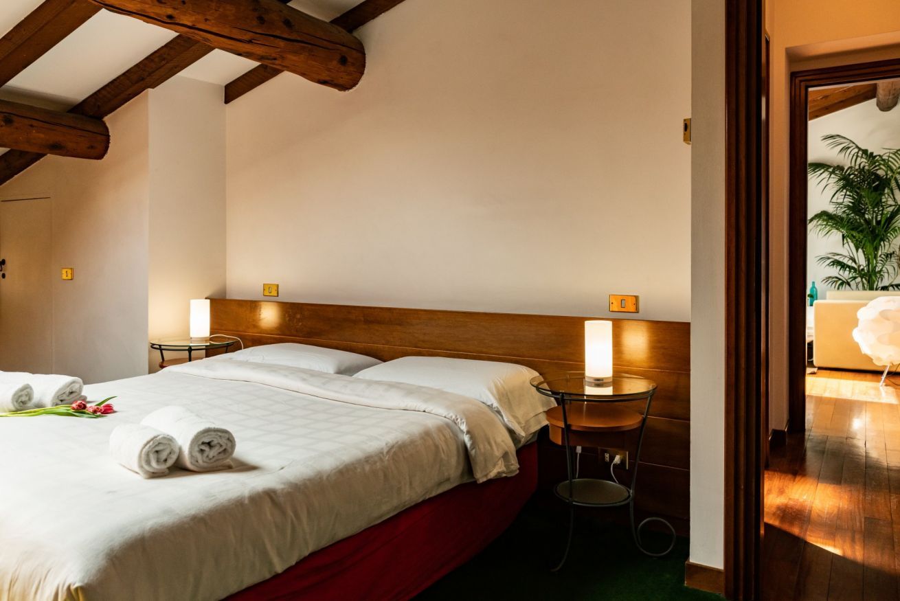 Habitación de hotel con techo de madera, una cama con sábanas blancas, dos mesitas de noche con lámparas y vistas a una planta a través de una puerta abierta.