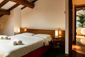 Een hotelkamer met een houten plafond, een bed met witte lakens, twee nachtkastjes met lampen, en uitzicht op een plant door een geopende deur.
