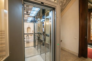 Un ascensor con puerta de vidrio, un botón verde y vistas a un vestíbulo con suelo de baldosas y un marco de puerta de madera.
