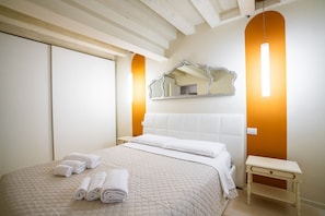 Una cama bien hecha con toallas enrolladas, una mesita de noche, un espejo y una lámpara de pared.