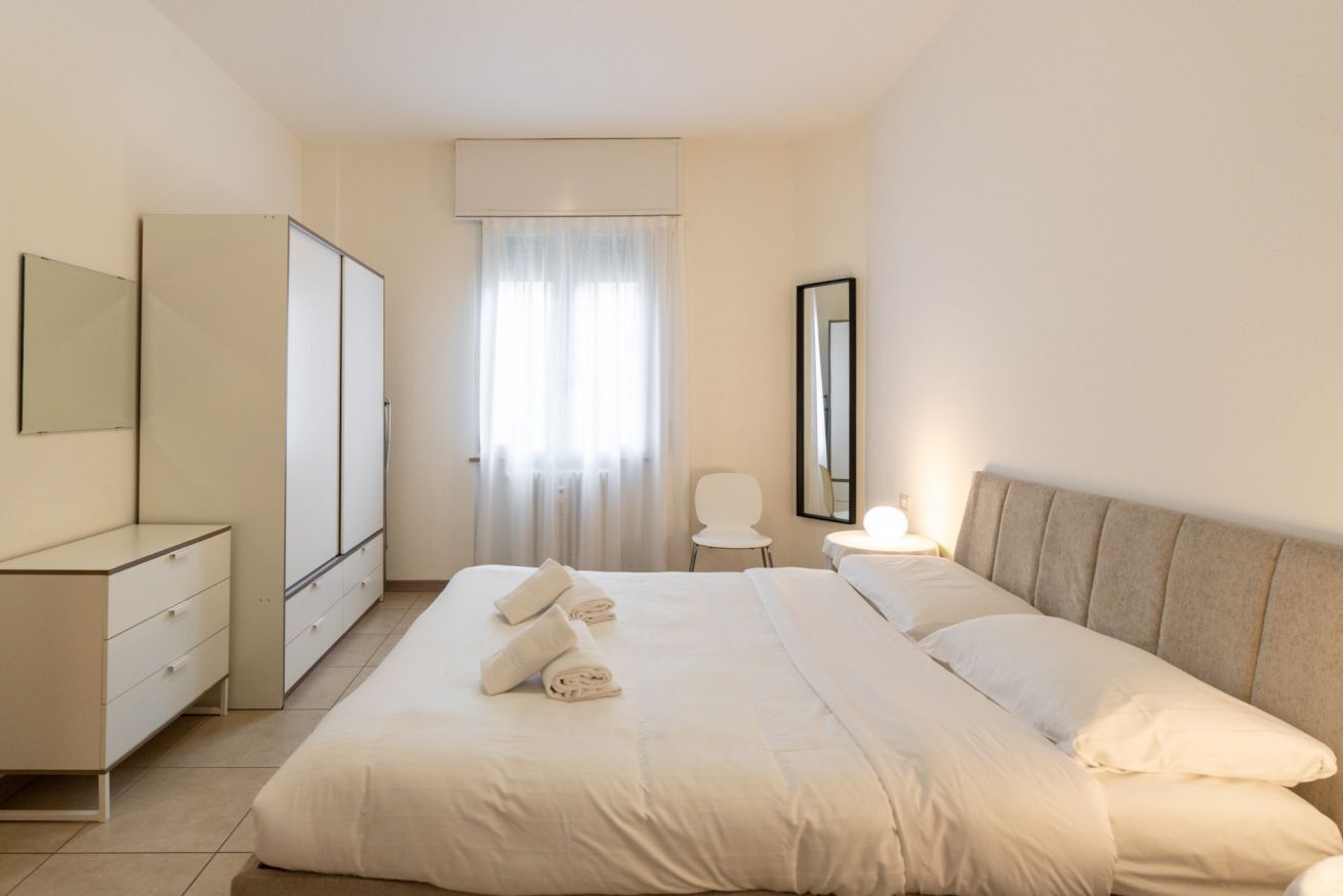 Una camera da letto con un letto, una cassettiera, uno specchio, una sedia e una finestra con tende.