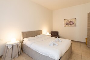 Un lit bien fait, avec du linge de lit blanc et deux oreillers, une table de chevet avec une lampe, une chaise et une œuvre d’art florale accrochée au mur.