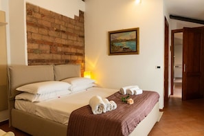 A neatly made bed with white and brown linens, a brick wall, and a painting of a landscape. - Magnificent Residence overlooking Piazza Erbe (Verona)