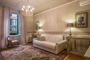 Una habitación con un sofá blanco, una alfombra estampada, un candelabro y una ventana con cortinas.