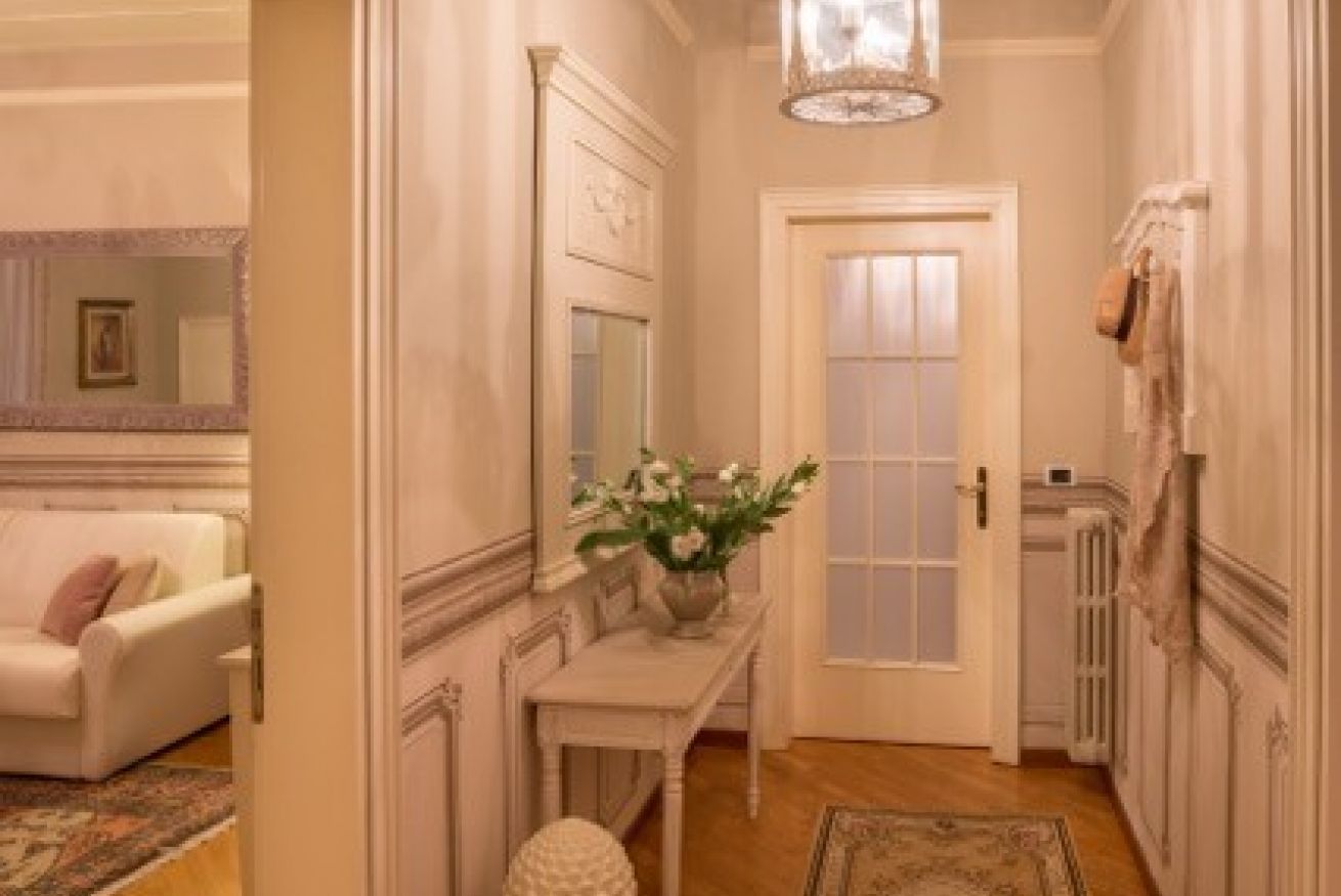A hallway with a white door, a white bench, a vase with flowers, and a chandelier.