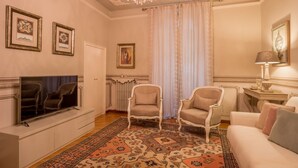 Een woonkamer met een televisie, twee fauteuils, een bank, een kroonluchter en een tapijt met een patroon.