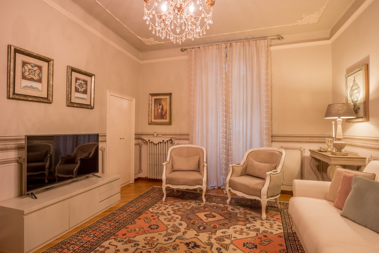 Un salon comprenant une télévision, deux fauteuils, un canapé, un lustre et un tapis à motifs.